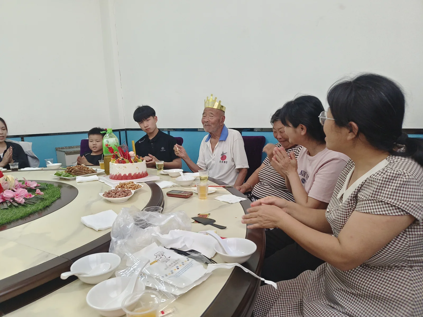 姥爷戴着生日冠坐在蛋糕旁，家人围坐拍手